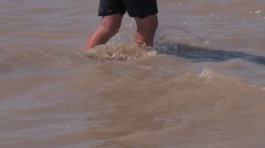 Man wearing swimming shirts and carring wakeboard is walking in the dirty water flow. Legs closeup.