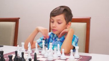 Mavi kareli gömlekli çocuk satranç oynar. Arkadaki ışık duvarı, yakın plan, tahta satranç tahtası..