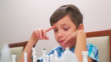 Mavi kareli gömlekli çocuk satranç oynar. Arkadaki ışık duvarı, yakın plan, tahta satranç tahtası..
