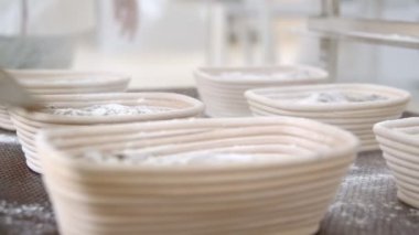 Fırıncılar, masada duran bir sürü hazır ekmeğin üzerine plastik bir kepçeden un serperler. Masada elektronik teraziler de var..