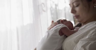 Beyaz tişört giyen anne bebeği elinde tutuyor. - Yakın çekim. Kapalı çekim. Arkasında beyaz perdeler olan pencere.