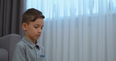 Odada gri tişört giyen bir çocuk var. Orta mesafeden. Arkasında gri, gri ve beyaz perdeler var..