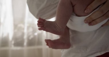 Beyaz tişört giyen anne bebeği elinde tutuyor. - Yakın çekim. Kapalı çekim. Arkasında beyaz perdeler olan pencere.