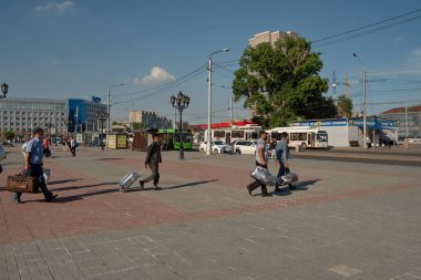 Krasnoyarsk, Krasnoyarsk Bölgesi, RF - 11 Temmuz 2021: Güney cumhuriyetlerinden göçmenler güneşli bir yaz gününde bavullarıyla istasyon meydanındaki otobüs durağına yürüyorlar.