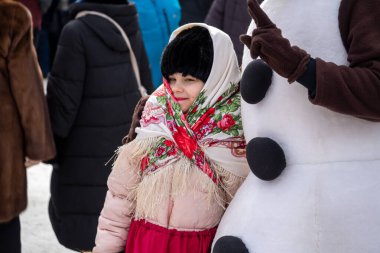 Krasnoyarsk, Krasnoyarsk Bölgesi, RF - 14 Mart 2021: Rus türbanlı bir kız, bir şehir parkındaki Farewell to Winter festivalinde bir kardan adam oyuncusunun yanında duruyor.