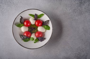 Düz İtalyan caprese salatası, olgun domates, taze fesleğen ve gri arka planda beyaz tabakta mozzarella peyniri.