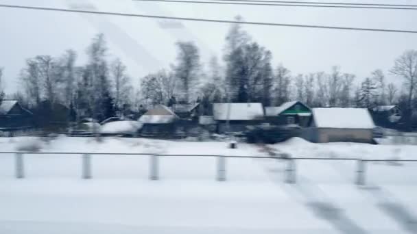 Θέα από το παράθυρο του κινούμενου τρένου σε χιονισμένο χωριό μονώροφα σπίτια, φράχτη, δέντρα σε συννεφιασμένη μέρα — Αρχείο Βίντεο