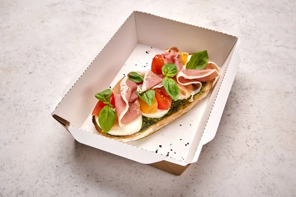 Bruschetta with mozzarella, Parma ham and pesto in white cardboard container on light background — Stock Photo, Image