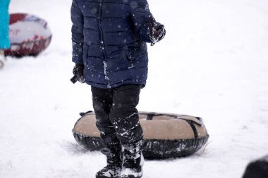 Koyu renk tulumlu küçük bir çocuk buz kaydırağından kayıyor, kış çocuk oyunları, kışın parkta bir çocuk. Oğlan tüple kaydırağa tırmandı.