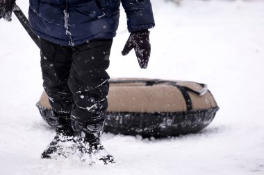 Koyu renk tulumlu küçük bir çocuk buz kaydırağından kayıyor, kış çocuk oyunları, kışın parkta bir çocuk. Oğlan tüple kaydırağa tırmandı.