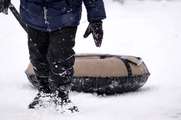 Koyu renk tulumlu küçük bir çocuk buz kaydırağından kayıyor, kış çocuk oyunları, kışın parkta bir çocuk. Oğlan tüple kaydırağa tırmandı.
