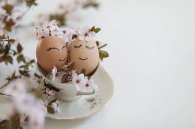 Creative easter eggs with cute face, sleepy eyes and spring flowers in tea cup on white background. Happy Easter concept, mockup, copy space. 