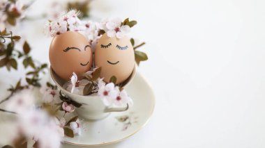 Creative easter eggs with cute face, sleepy eyes and spring flowers in tea cup on white background. Happy Easter concept, mockup, copy space. 