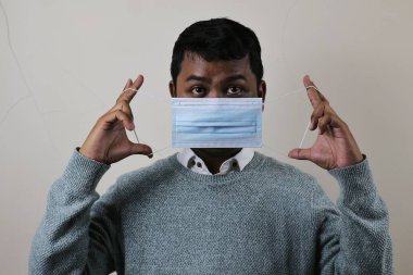 Genç Asyalı bir adam maskeyi doğru bir şekilde nasıl takacağını gösteriyor, maskenin kayışını tutuyor ve geriyor.