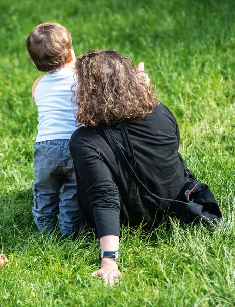 Mujer sentada en la hierba señalando algo a su hijo muy joven de pie junto a ella — Foto de Stock