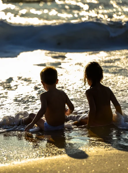 Duas crianças pequenas sentadas nas ondas do oceano sob um belo pôr do sol — Fotografia de Stock