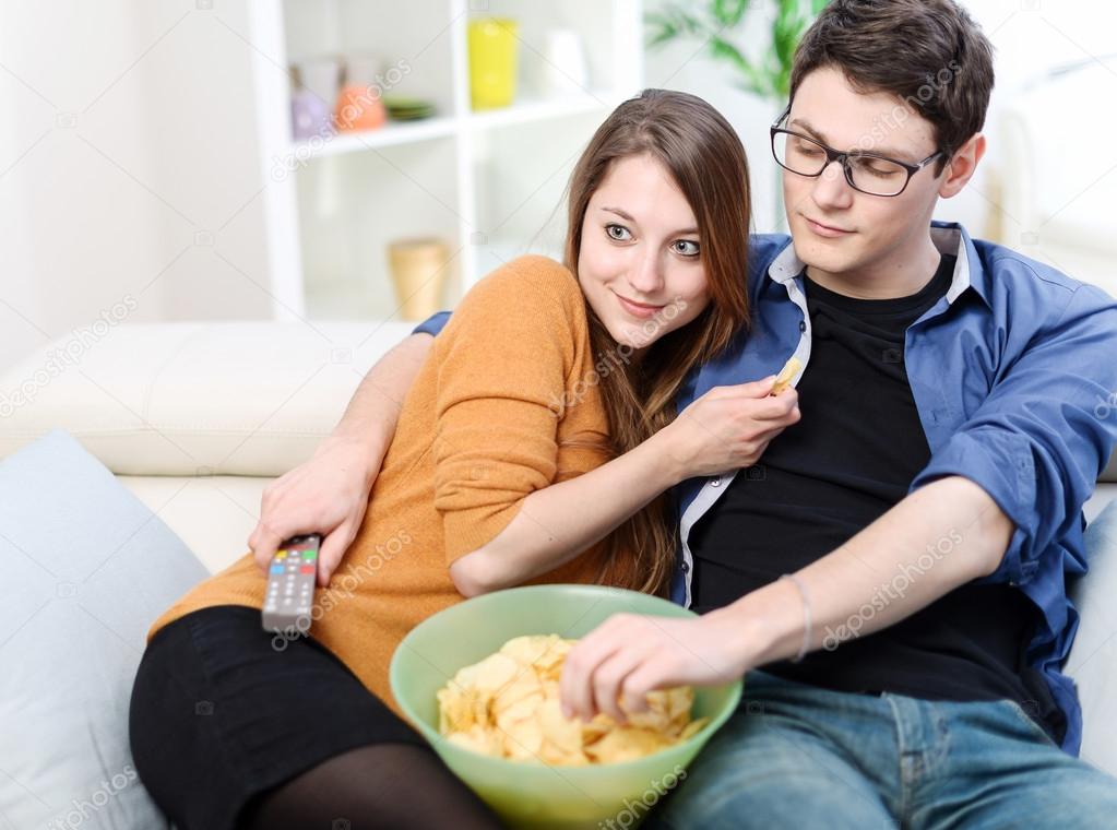 Beautiful young married looking for tv and eating on a sofa at apartment