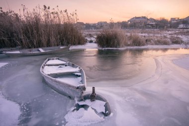Donmuş nehir. Kışın nehir kıyısında kar ve buzun altında eski ahşap bir tekne. Güzel kış gündoğumu.