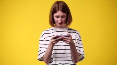Sarı arka planda izole edilmiş telefonundan bir şey izleyen hoş bir kadının 4K videosu. Teknoloji kavramı.
