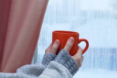 An orange cup in the hands of a Caucasian woman. Against the background of frozen glass on the window in the room.