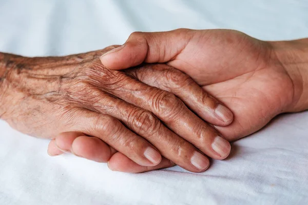 Hands of the old man and a man hand on the white bed in a hospital