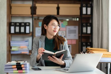 Asyalı bir kadın büroda oturmuş başka tarafa bakarken elinde kağıt bir kitap tutuyor. 