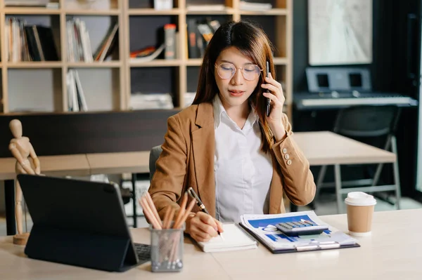 Asyalı iş kadını telefonda konuşuyor ve notlar alıyor, modern ofis, dizüstü bilgisayarlı masa, kahve ve belgeler. 