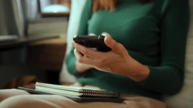 Yeşil kazaklı bir kadın, evdeki koltukta oturuyor ve cep telefonu kullanıyor. 