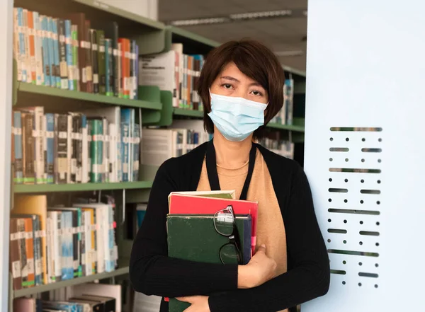 Woman with face mask holding hard cover books with her arm,between prepare searching data,at library