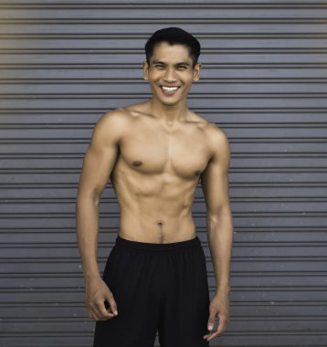 Gülümseyen yüzlü genç adam, arka planda duruyor, egzersizden dolayı kaslı ve kaslı, bulanık ışık etrafında.