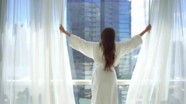 Otel odasında perdeleri açan bornozlu güzel Asyalı kadının görüntüleri.