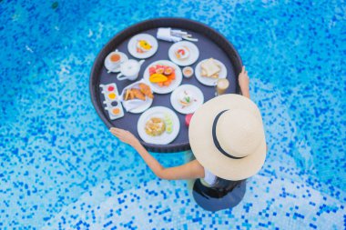 Asyalı güzel bir kadının portresi yüzme havuzunda yüzen kahvaltı tepsisiyle mutlu bir gülümseme