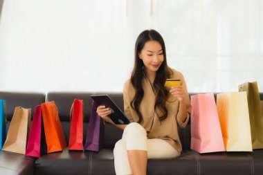 Güzel Asyalı bir kadının portresi. İnternetten alışveriş yapıyor. Kredi kartı ve akıllı telefonuyla oturma odasındaki alışveriş çantasının etrafında.