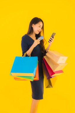 Portre güzel genç Asyalı kadın alışveriş çantası kredi kartı ve akıllı cep telefonu sarısı izole bir arka plan.