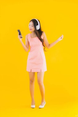 Güzel bir Asyalı kadın portresi. Sarı izole edilmiş arka planda akıllı cep telefonu ve kulaklıkla müzik dinliyor.