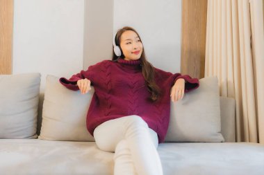 Güzel bir Asyalı kadının portresi oturma odasındaki kanepede müzik dinlemek için kulaklık kullanıyor.