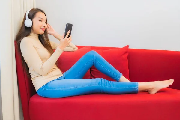 肖像画美しい若いアジアの女性はソファの上で音楽を聴くためのヘッドフォンとスマートフォンを使用します — ストック写真