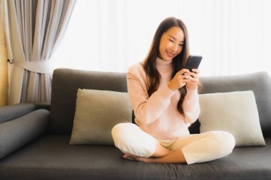 Oturma odasındaki koltukta akıllı cep telefonu kullanan güzel Asyalı kadın portresi.