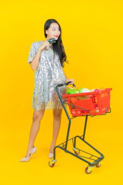 Asyalı güzel bir kadının portresi. Sarı arka planda süpermarketten alınmış alışveriş sepetiyle gülümsüyor.