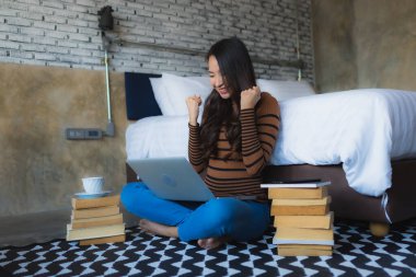 Genç Asyalı kadın yatak odasının içinde elinde kahve fincanı ve kitapla dizüstü bilgisayar kullanıyor.