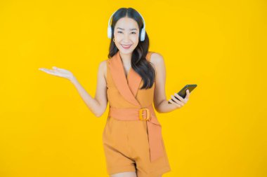 Sarı arka planda müzik dinlemek için kulaklığı ve akıllı telefonu olan güzel Asyalı kadın portresi.