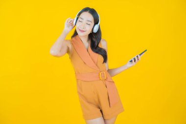 Sarı arka planda müzik dinlemek için kulaklığı ve akıllı telefonu olan güzel Asyalı kadın portresi.