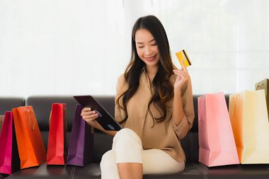 Güzel Asyalı bir kadının portresi. İnternetten alışveriş yapıyor. Kredi kartı ve akıllı telefonuyla oturma odasındaki alışveriş çantasının etrafında.