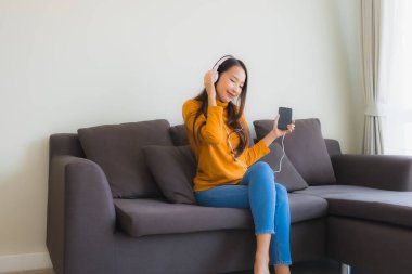 Genç Asyalı kadın oturma odasındaki kanepede müzik dinlemek için akıllı cep telefonu ve kulaklık kullanıyor.