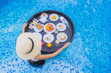 Asyalı güzel bir kadının portresi yüzme havuzunda yüzen kahvaltı tepsisiyle mutlu bir gülümseme