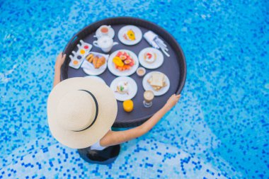 Asyalı güzel bir kadının portresi yüzme havuzunda yüzen kahvaltı tepsisiyle mutlu bir gülümseme