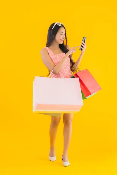 亚洲年轻貌美的女子 头戴五颜六色的购物袋 手持智能手机 背景为黄色 背景为孤立的信用卡 — 图库照片
