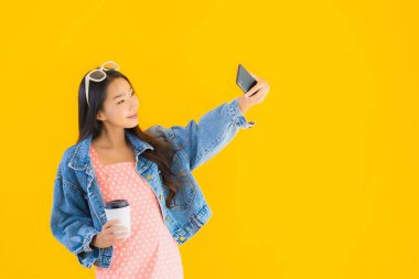 Kahve fincanı ve sarı izole edilmiş arka planda akıllı cep telefonlu güzel Asyalı kadın portresi.