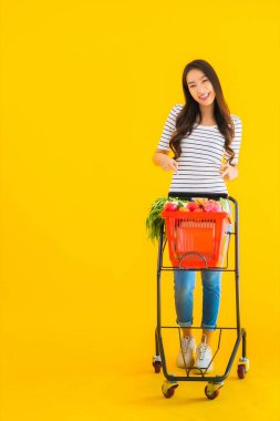 Güzel genç Asyalı kadın süpermarketten alışveriş yapıyor ve sarı izole arka planda el arabası var.