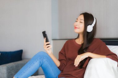 Güzel bir Asyalı kadın yatak odasındaki yatak odasında müzik dinlemek için kulaklıklı akıllı cep telefonu kullanıyor.